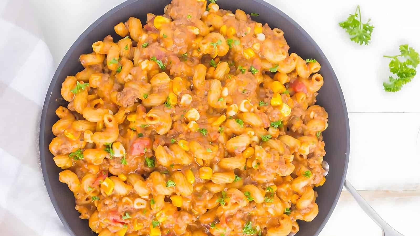 A skillet filled with pasta, ground meat, corn, and chopped tomatoes in a creamy sauce, garnished with parsley.