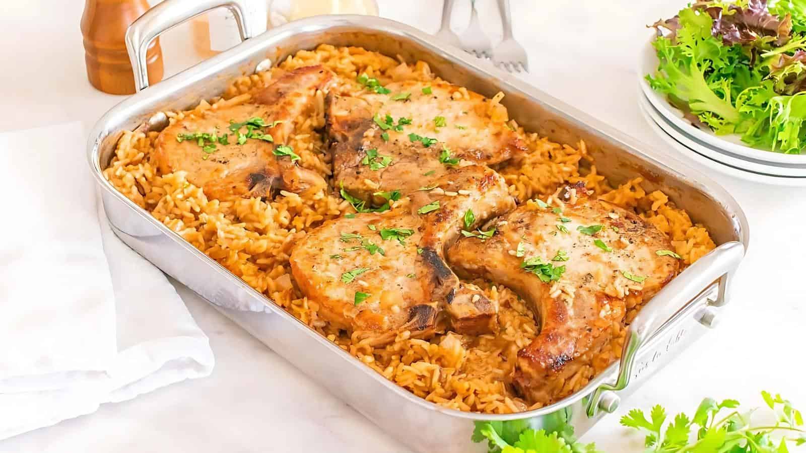 A baking dish filled with cooked pork chops on top of seasoned rice, garnished with chopped herbs, sits on a white surface next to a salad and plates.