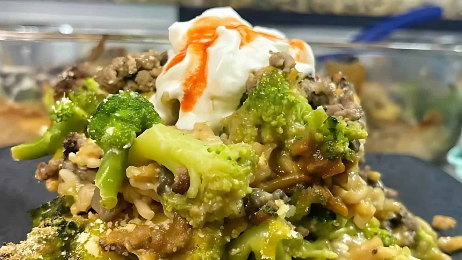 Close-up of a broccoli and rice casserole with ground meat, topped with a dollop of sour cream and a drizzle of hot sauce.