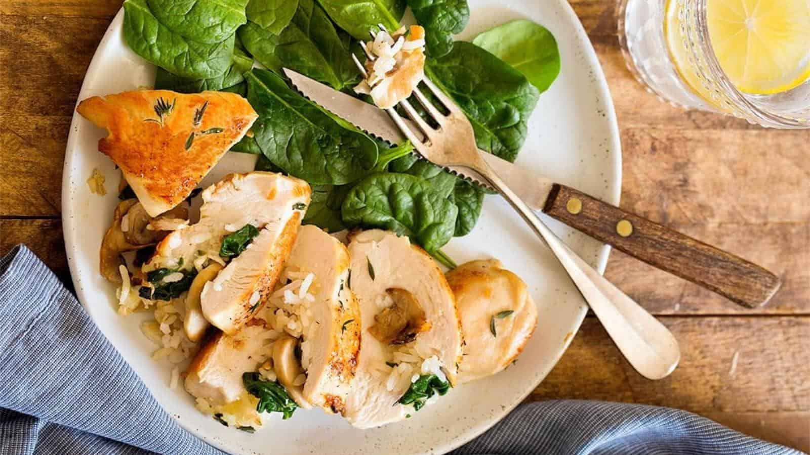 A plate with sliced stuffed chicken breast, fresh spinach leaves, a piece of bread, and a glass of water with a lemon slice on a wooden table.
