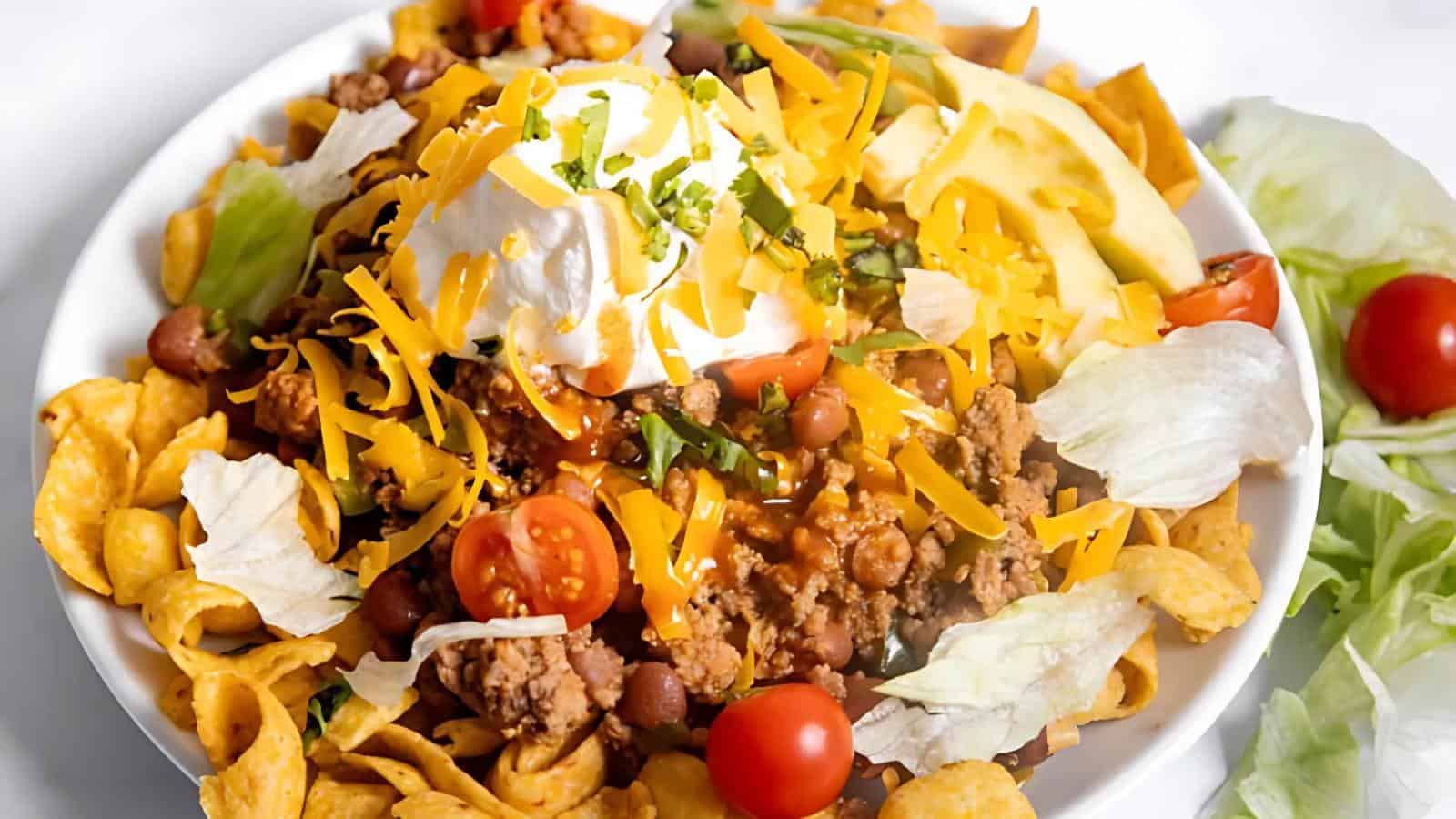 A plate of taco salad with corn chips, ground meat, beans, lettuce, cheese, cherry tomatoes, sour cream, and avocado slices.