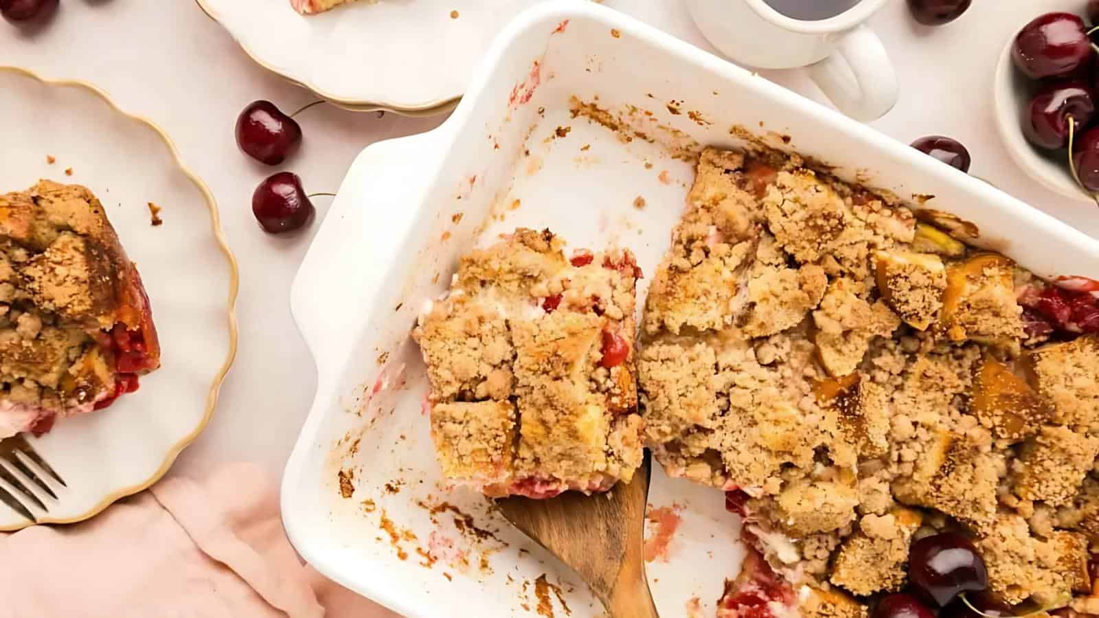 A partially eaten cherry cobbler in a baking dish, with a wooden spatula. Cherries and plates surround the dish on a light surface.