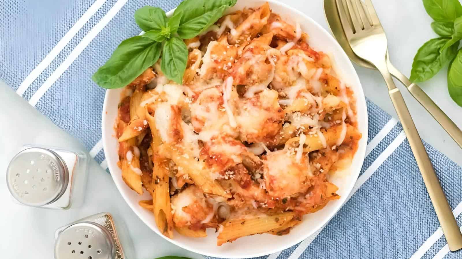 A bowl of baked penne pasta topped with melted cheese and tomato sauce, garnished with fresh basil, on a blue and white napkin with gold utensils and salt and pepper shakers nearby.