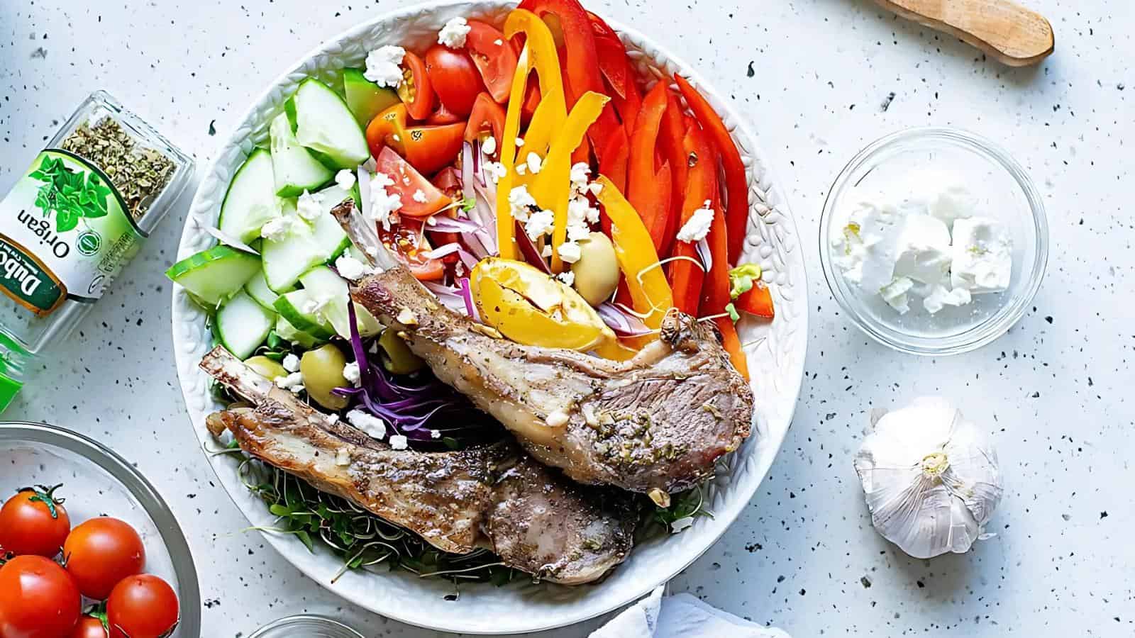 A plate with grilled lamb chops, sliced cucumber, tomatoes, red and yellow bell peppers, olives, red onion, feta cheese, and herbs, surrounded by ingredients on a white countertop.