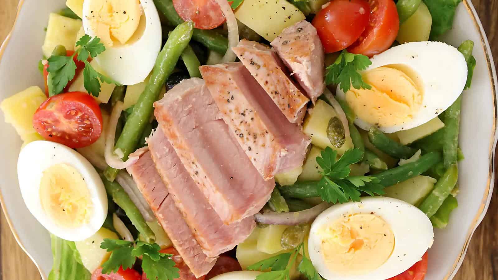 A plate of salad with sliced seared tuna, hard-boiled eggs, green beans, cherry tomatoes, potatoes, olives, onions, and parsley.
