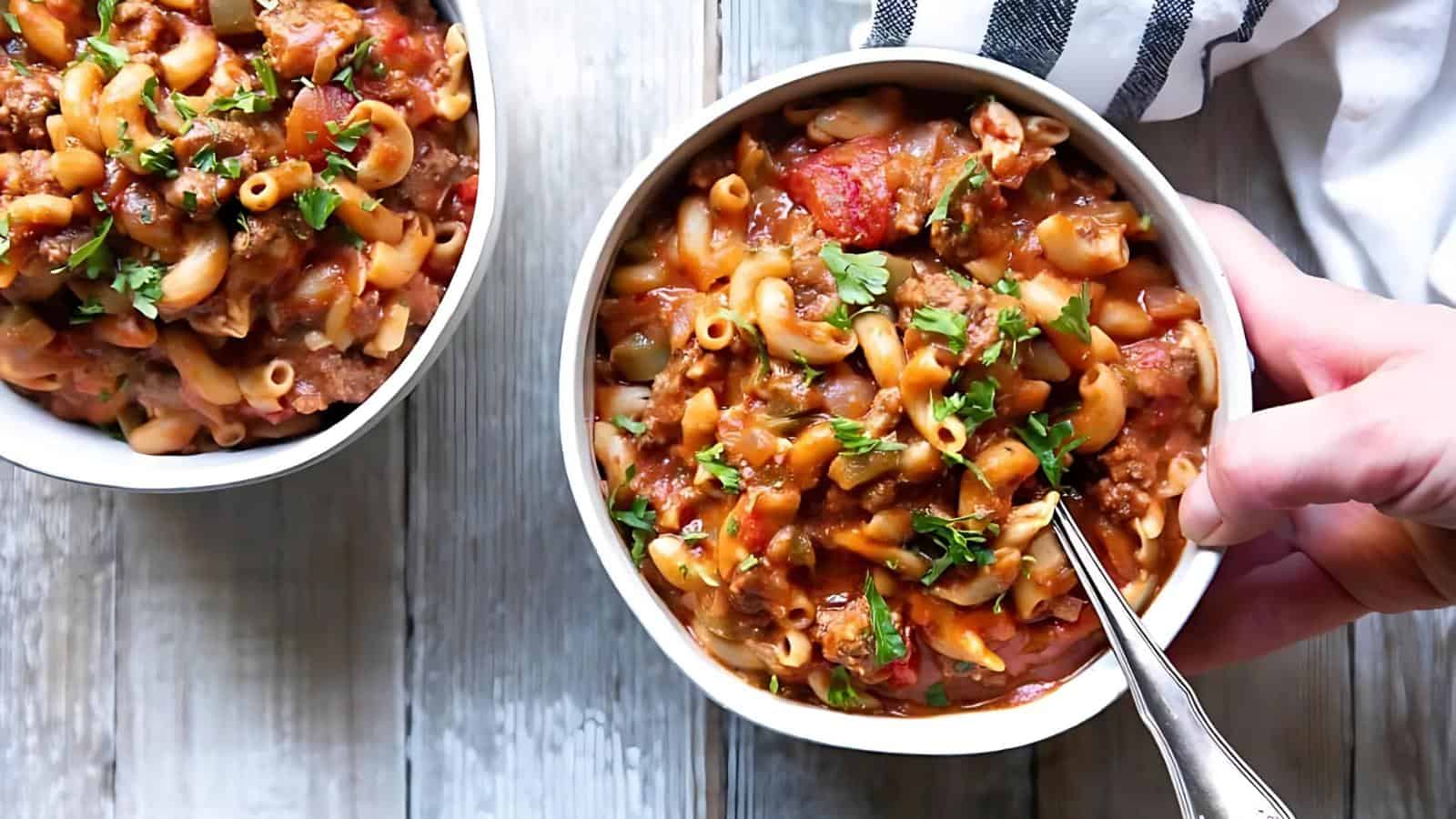 A hand holds a bowl of macaroni pasta with ground meat, tomato sauce, and herbs, with another similar bowl partially visible on a wooden surface.