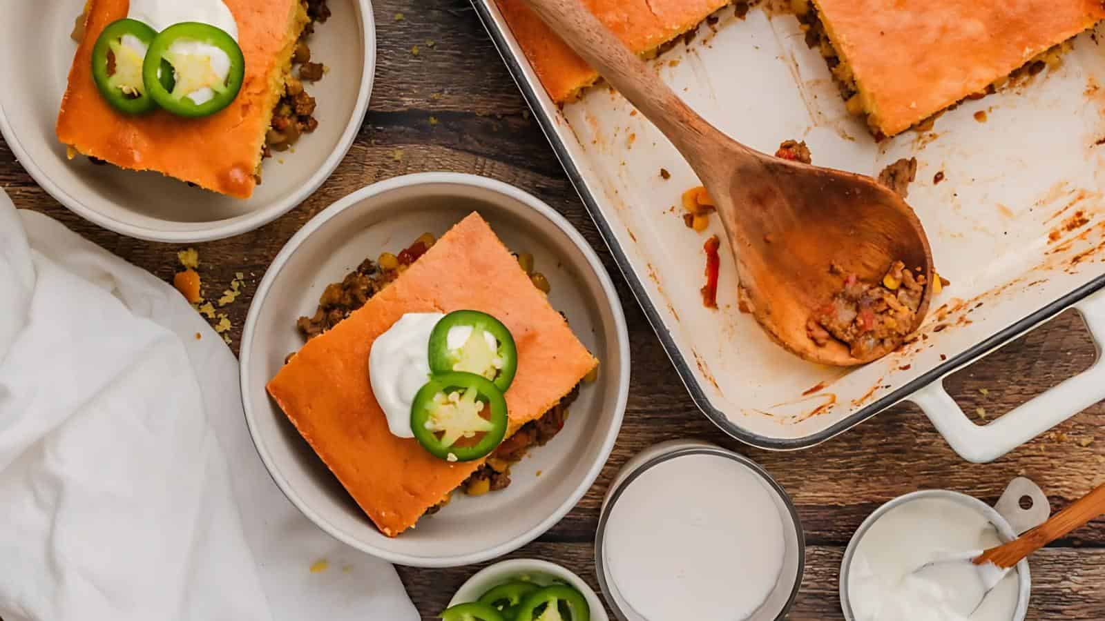Casserole slices with toppings on plates, wooden spoon in a baking dish, jalapeños and cream nearby on a wooden table.