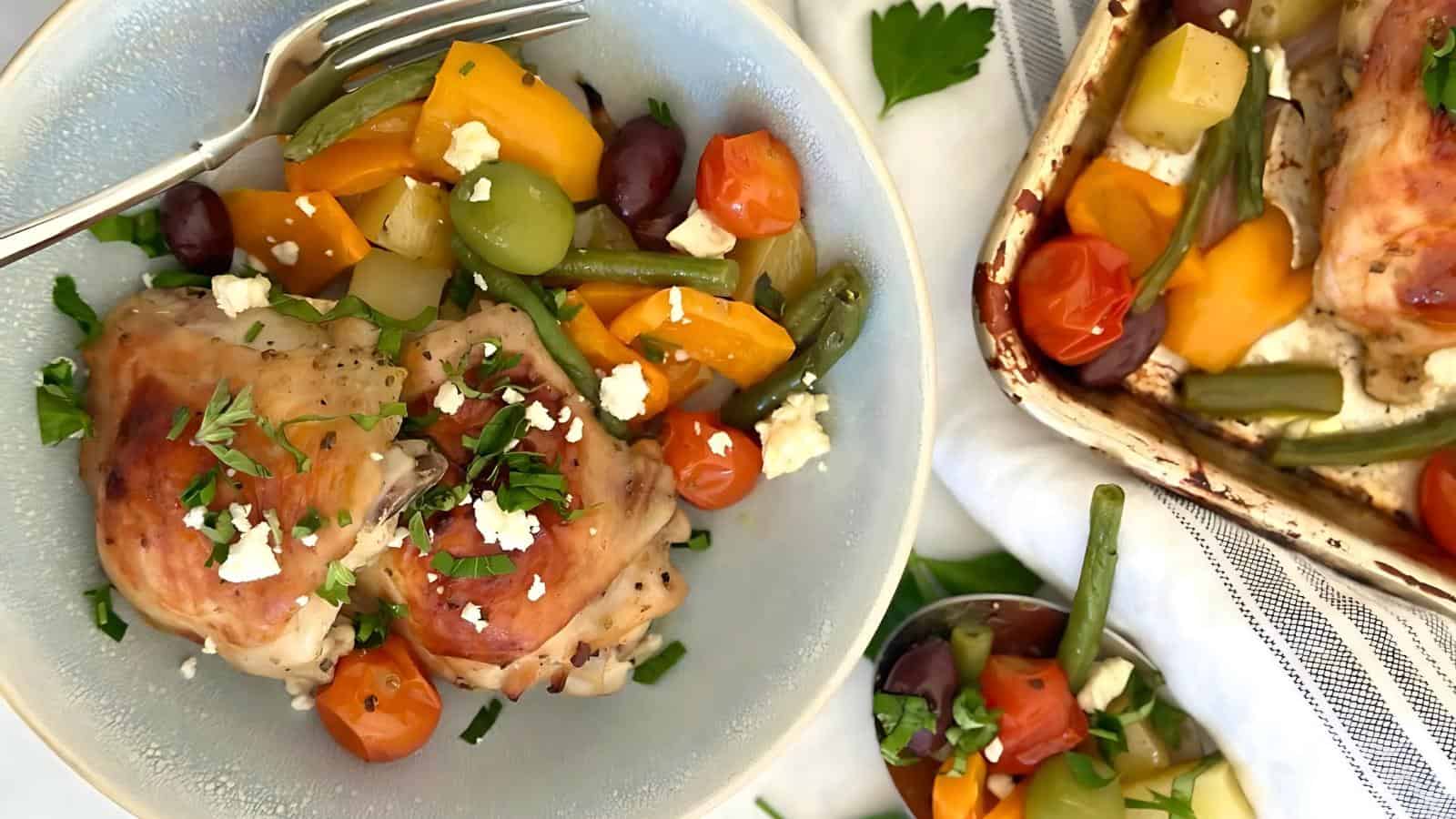 A plate with baked chicken thighs, mixed roasted vegetables, olives, and crumbled cheese, garnished with chopped parsley. A baking dish with the same food is nearby.