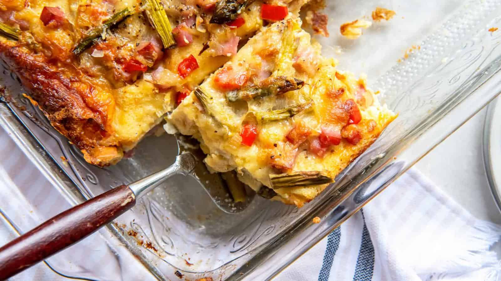 A slice of vegetable casserole with cheese, bell peppers, and asparagus in a glass baking dish, served with a metal spatula.