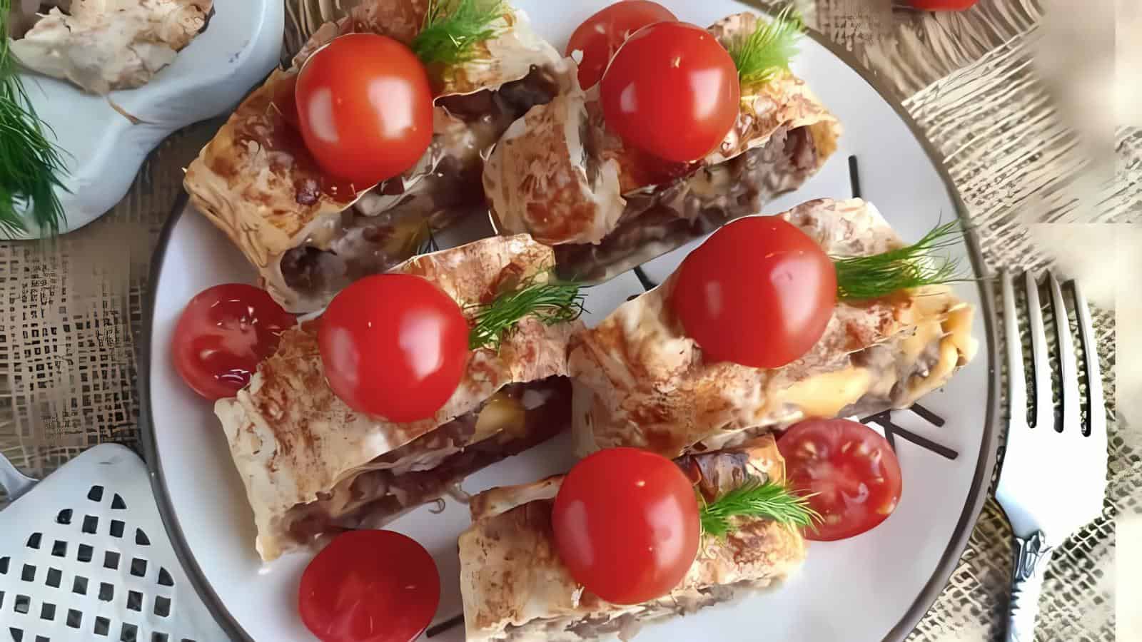 Five baked savory rolls filled with meat and topped with whole cherry tomatoes and dill, served on a white plate with extra cherry tomato halves on the side.