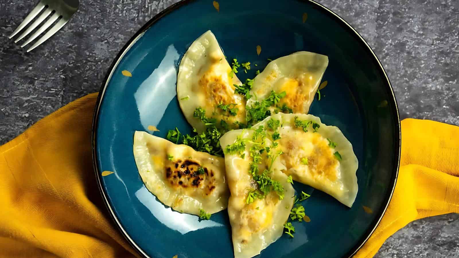Four cooked dumplings garnished with chopped herbs are arranged on a blue plate, set on a yellow cloth with a fork nearby.