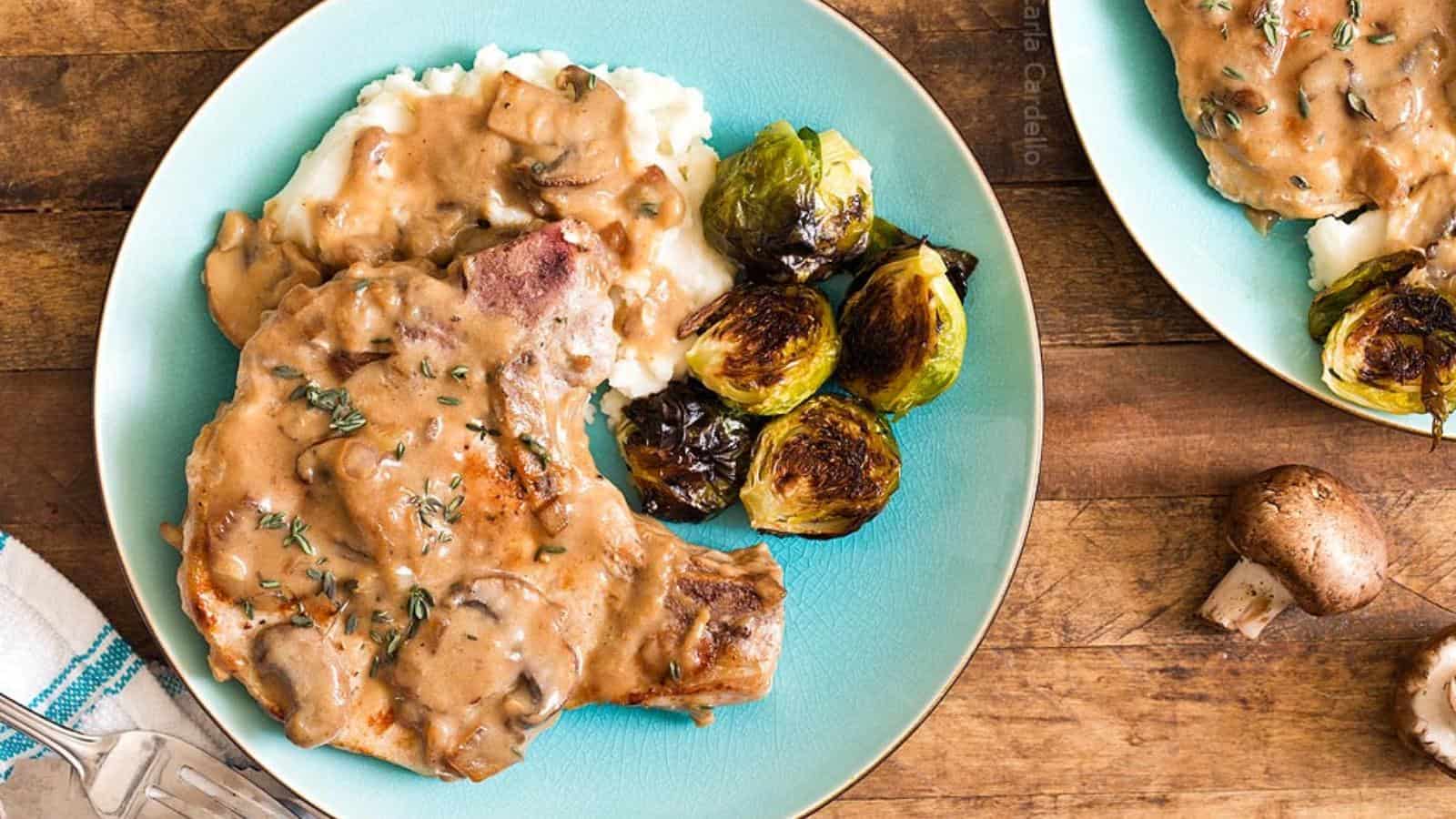 A plate with pork chops in mushroom gravy, mashed potatoes, and roasted Brussels sprouts sits on a wooden table.