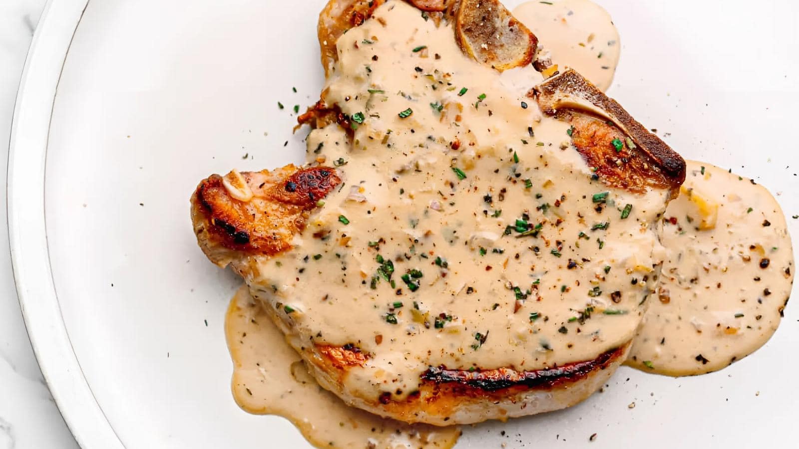 A cooked pork chop topped with creamy peppercorn sauce, garnished with herbs, served on a white plate.