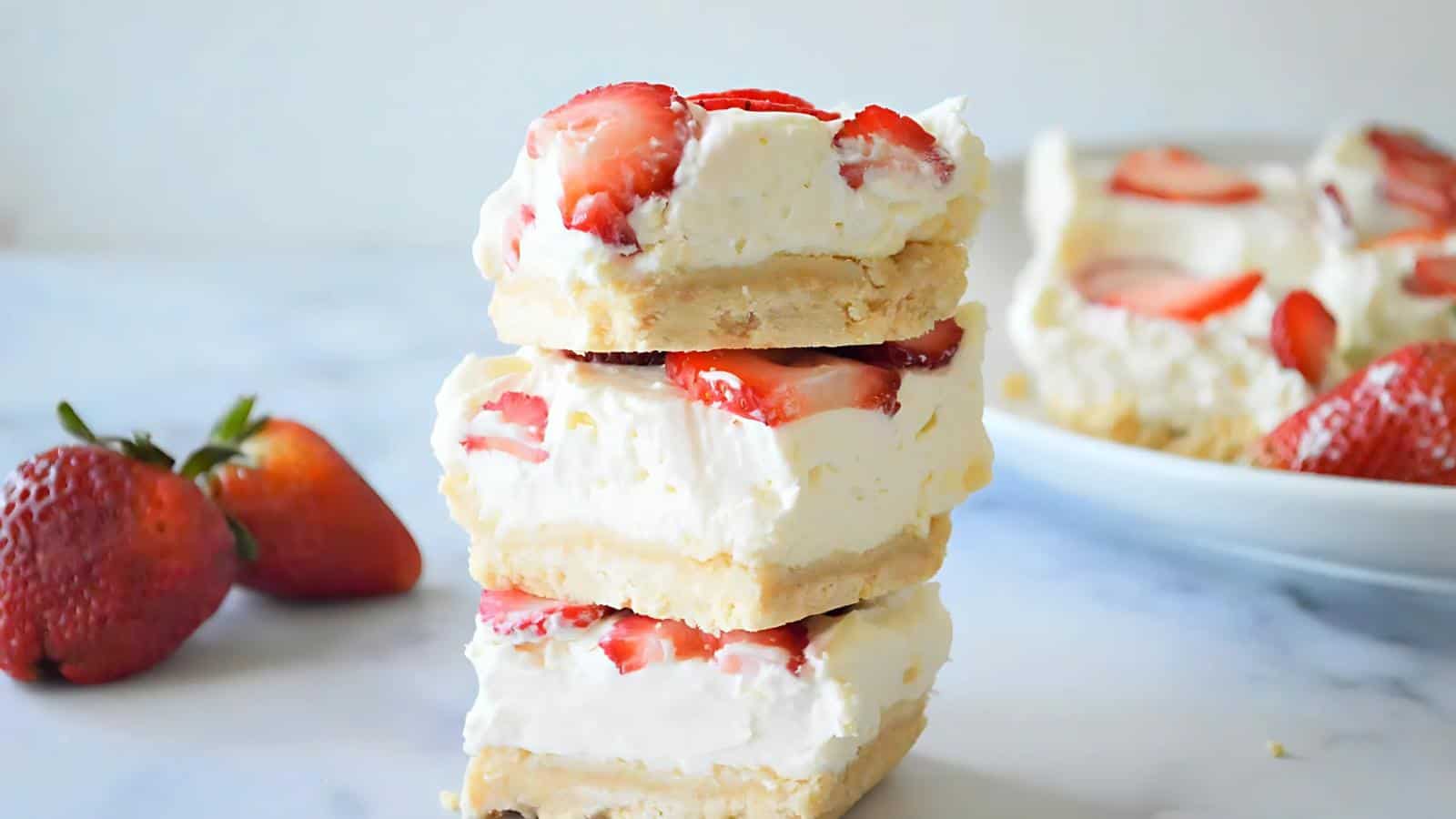 Three strawberry cheesecake bars stacked on top of each other, with fresh strawberries and a plate of bars in the background.