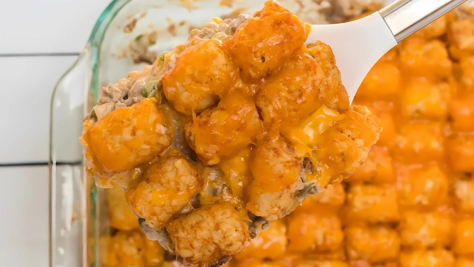 A serving spatula lifts a portion of tater tot casserole topped with melted cheddar cheese from a baking dish.