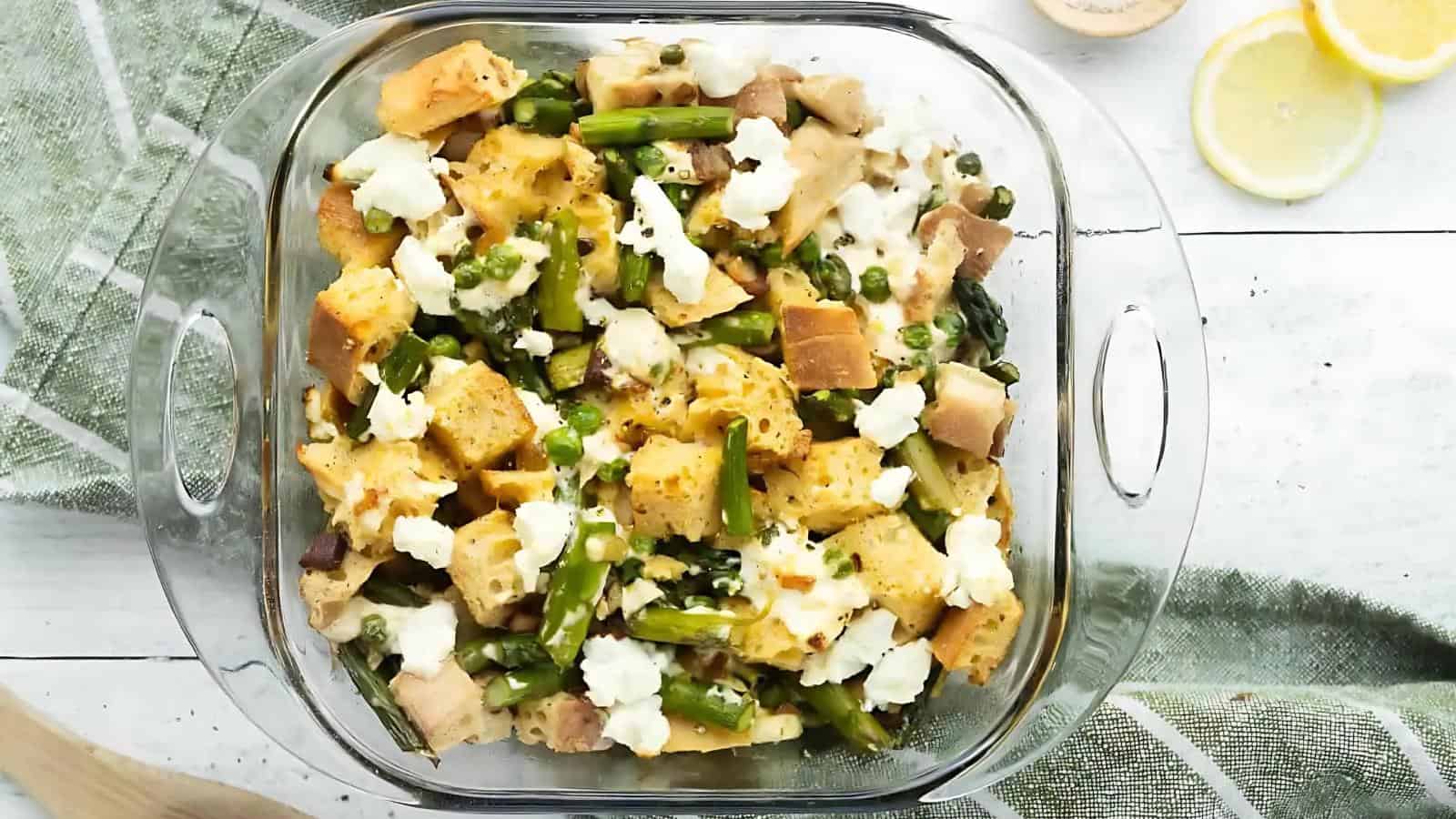 A glass baking dish filled with a casserole made of bread cubes, asparagus, green peas, and crumbled cheese.