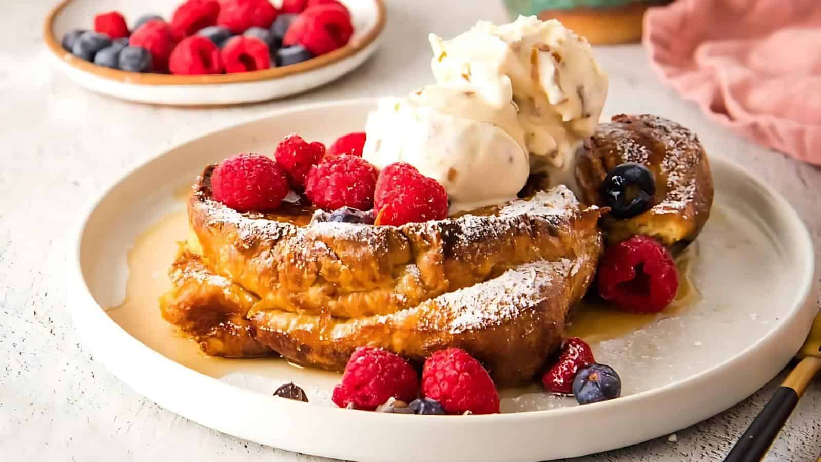 French toast topped with raspberries, blueberries, and a scoop of vanilla ice cream is on a plate, dusted with powdered sugar. Background includes a dish of raspberries.