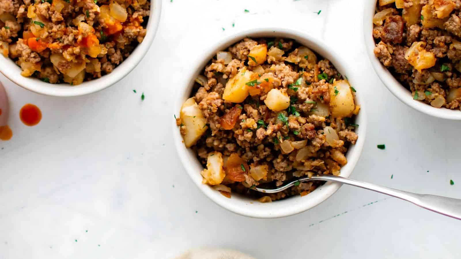A white bowl filled with ground beef, diced potatoes, onions, and vegetables, with a fork resting inside. Two similar bowls are partially visible nearby on a white surface.