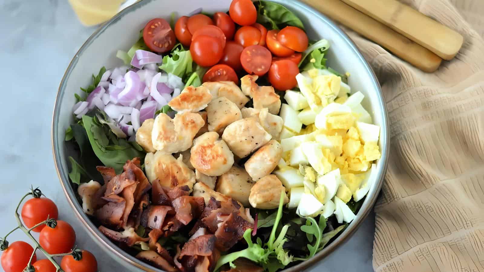 A bowl filled with greens, cherry tomatoes, chopped red onion, grilled chicken, hard-boiled eggs, and cooked bacon pieces, next to a beige napkin and utensils.