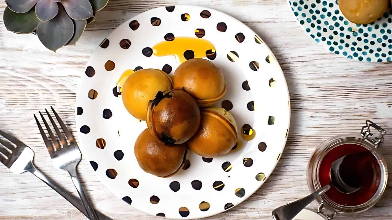 A white plate with black spots holds five round brown buns drizzled with yellow sauce, placed on a wooden table alongside utensils, a jar with red liquid, and a potted succulent.