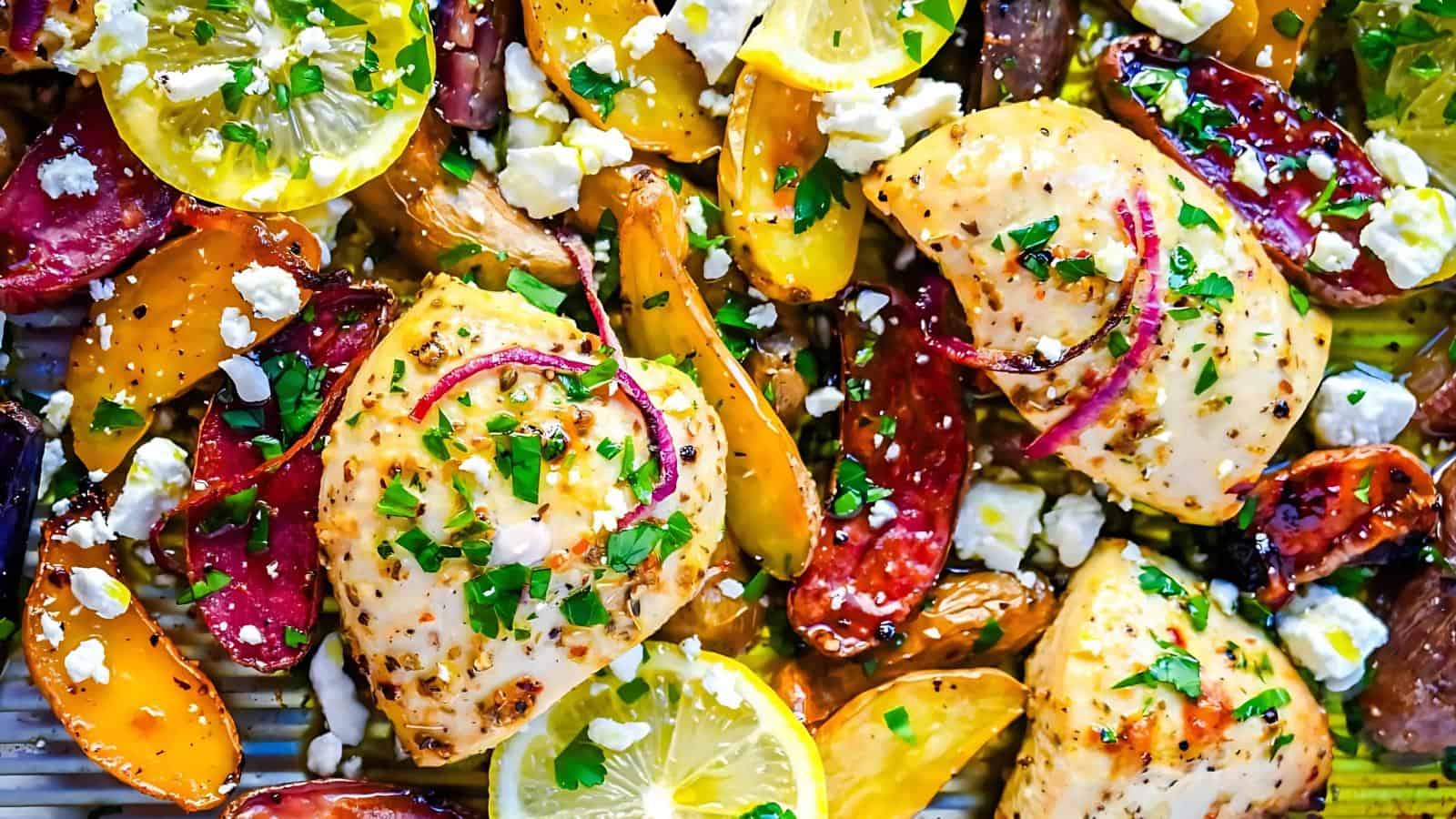 Sheet pan with baked chicken breasts, assorted roasted potatoes, red onions, lemon slices, feta cheese crumbles, and chopped herbs.