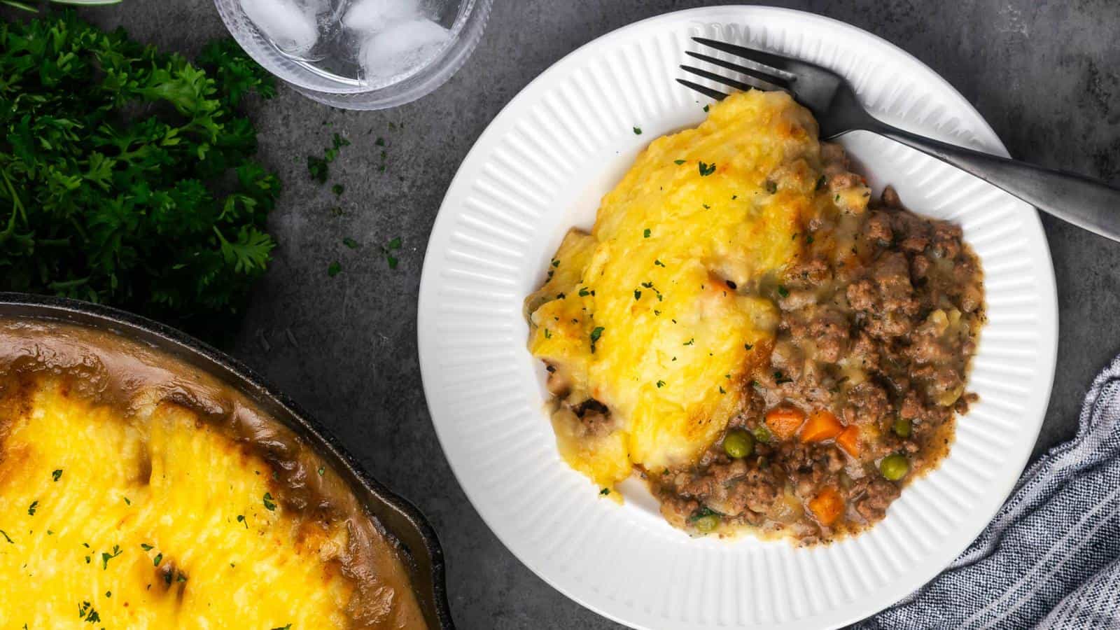 A plate of shepherd's pie with beef and vegetables beside a casserole dish containing more pie. A fork rests on the plate. Ice water and parsley sit nearby on a gray surface.
