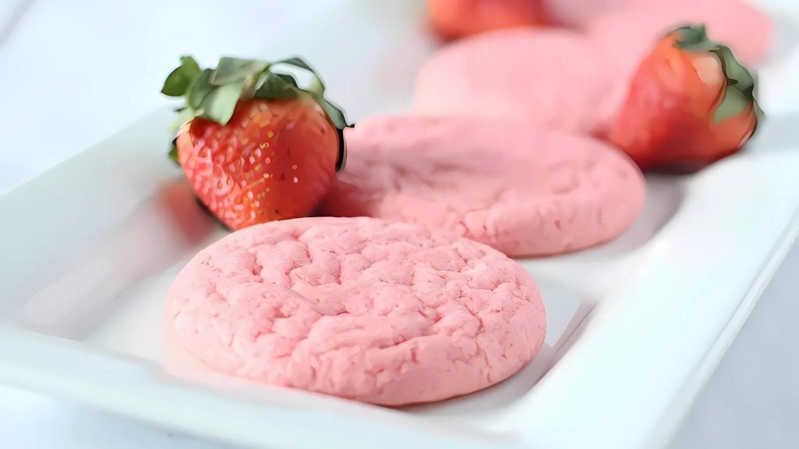 A white rectangular plate with three pink cookies and two whole strawberries arranged on it.