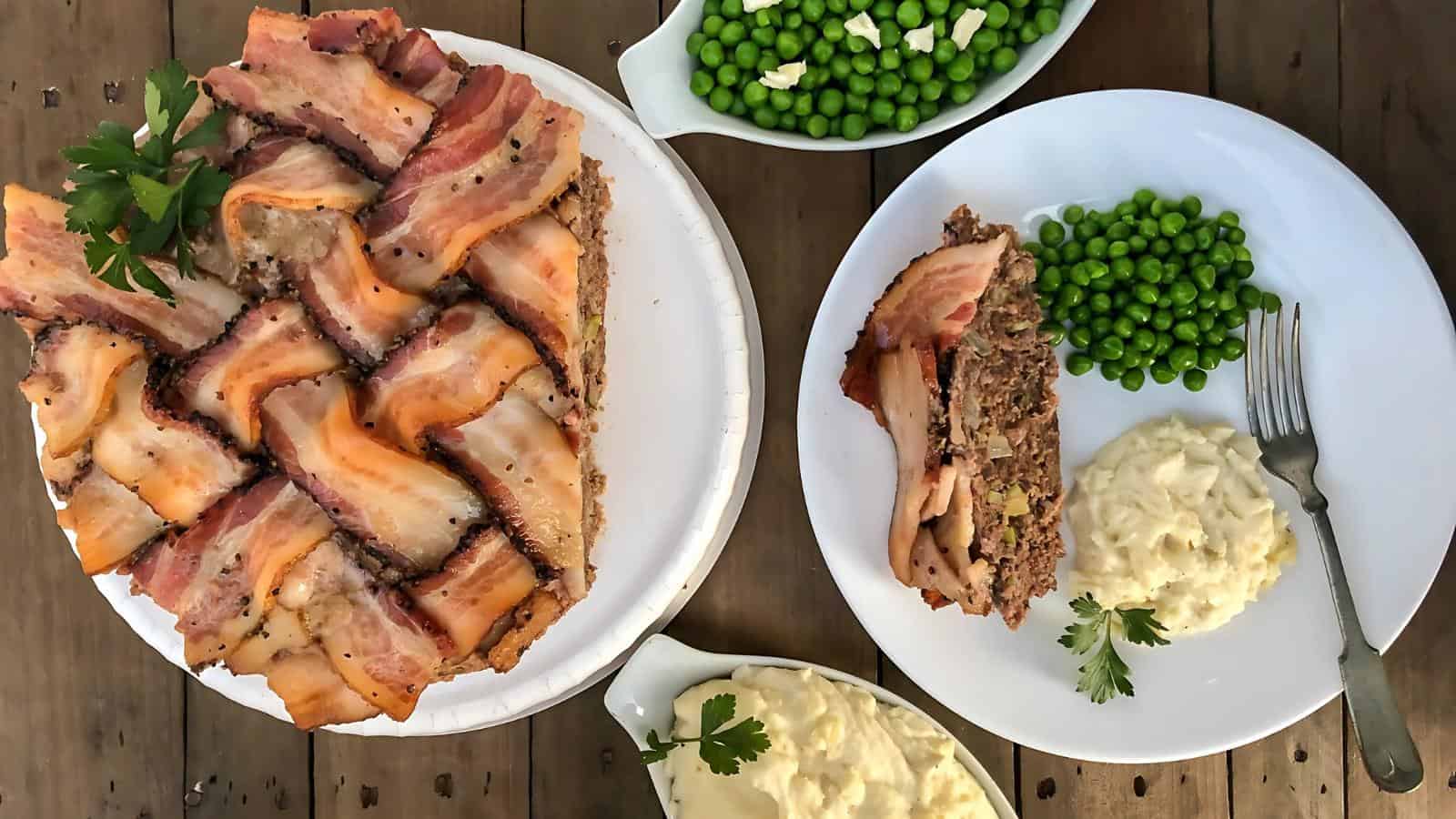 Bacon-wrapped meatloaf on a plate with peas and mashed potatoes. Whole meatloaf and additional sides in bowls are on a wooden table.