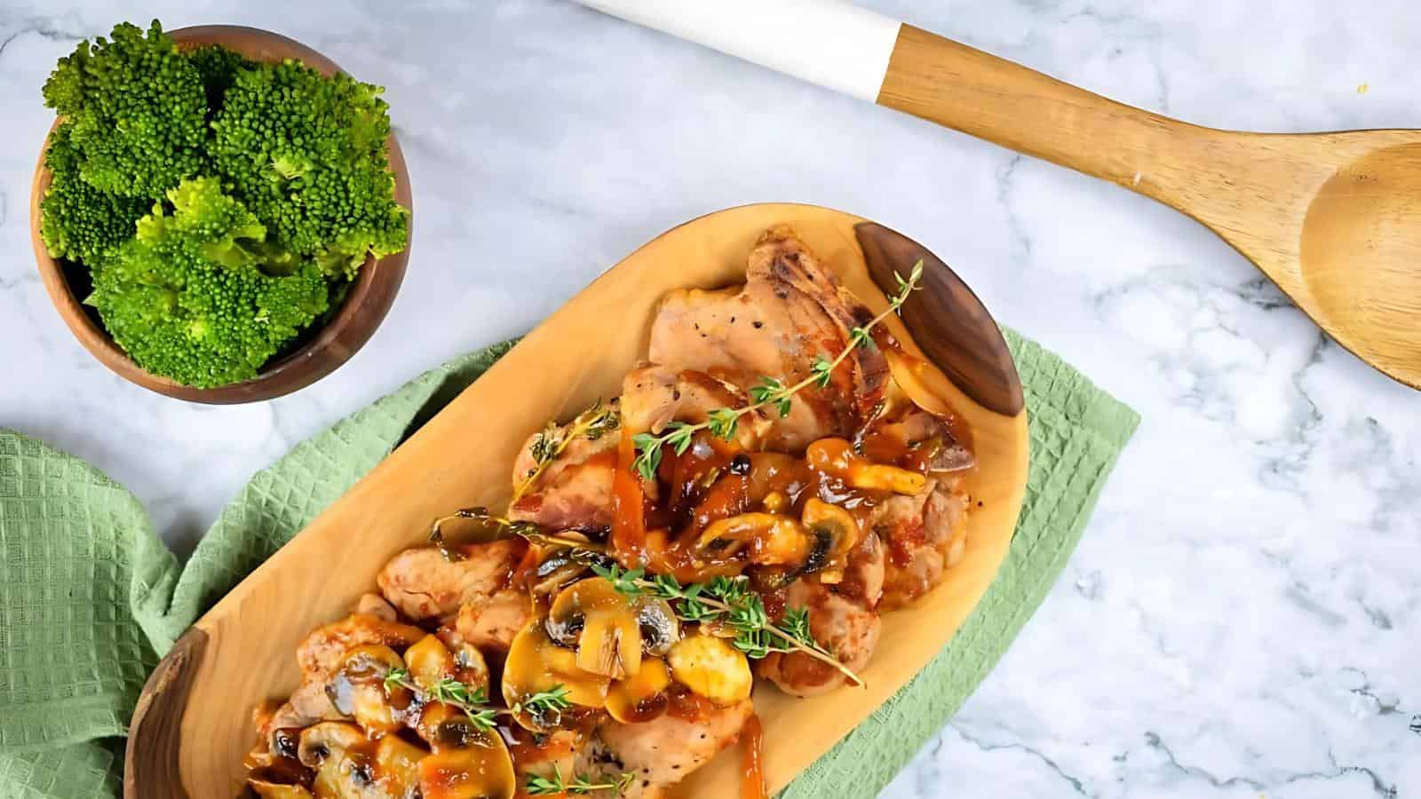 Wooden platter of cooked chicken with sauce and mushrooms, garnished with herbs, next to a bowl of broccoli, a green cloth, and a wooden spoon on a marble surface.