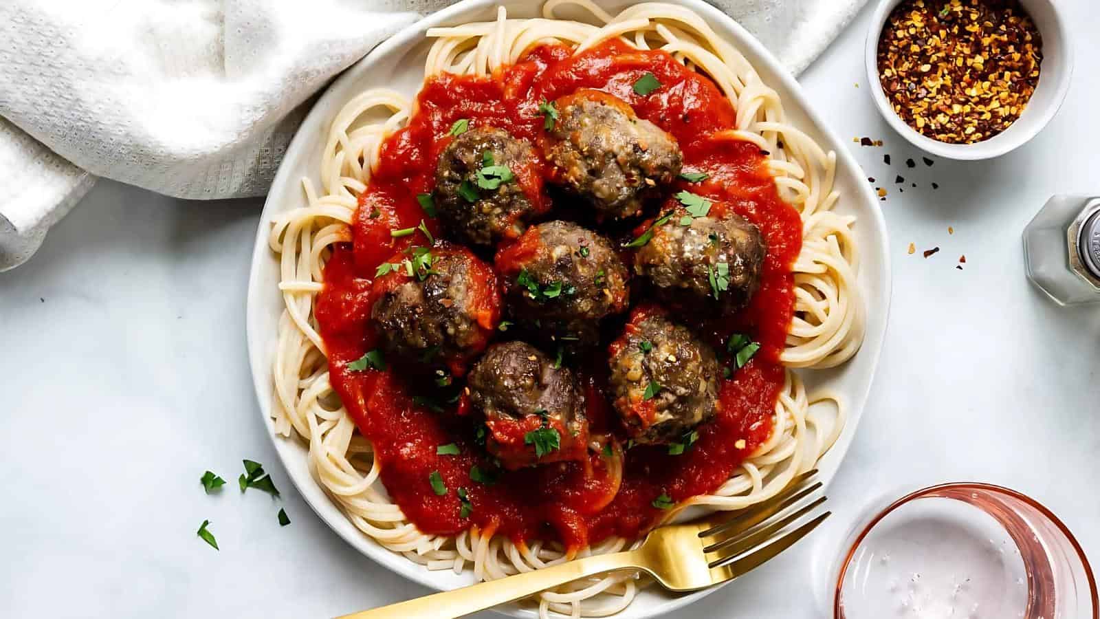 A plate of spaghetti topped with tomato sauce and meatballs, garnished with chopped parsley, beside a fork, a drink, and a small bowl of red pepper flakes.