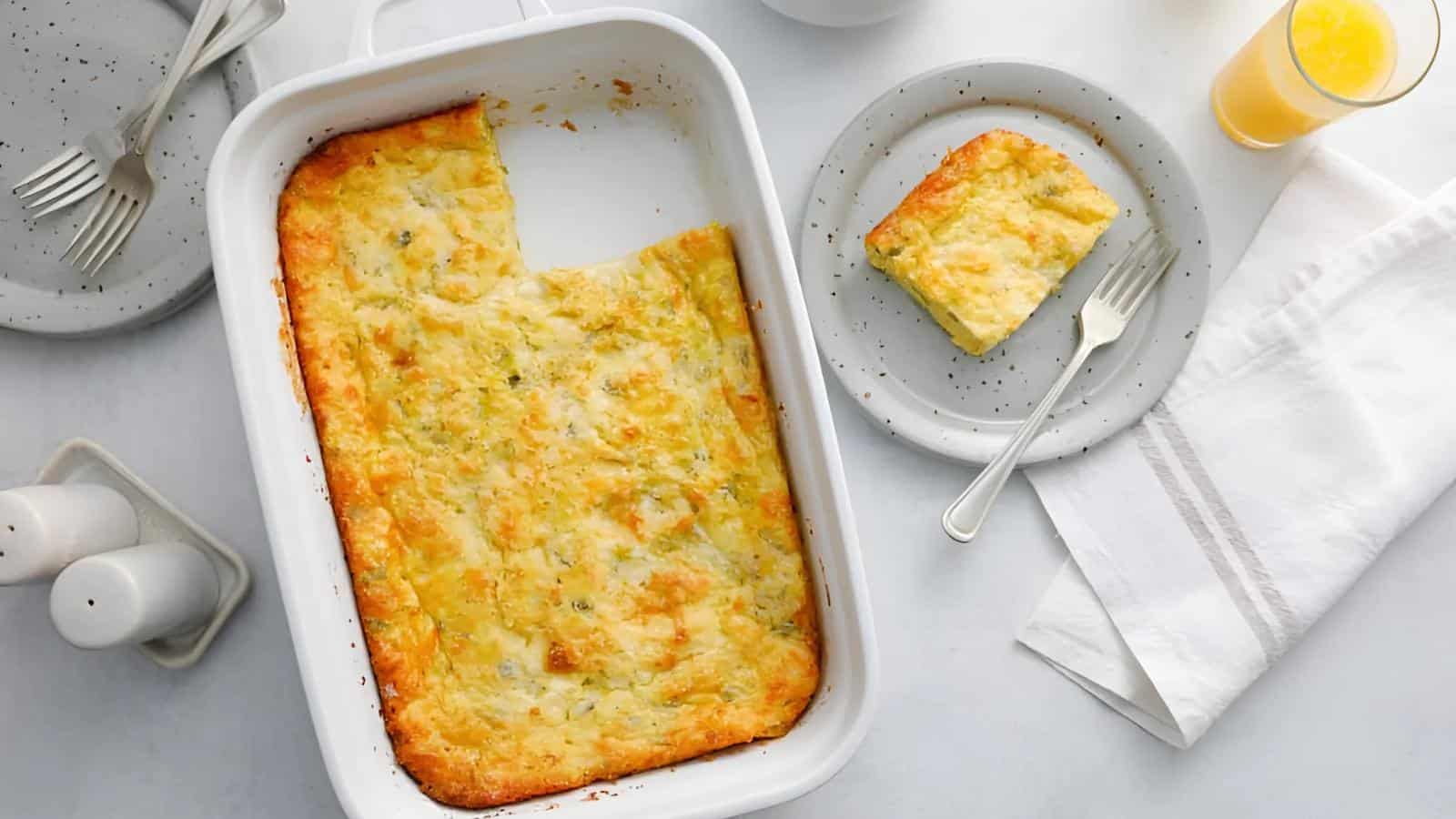 A baked casserole with a slice removed, placed on a plate beside it. There are salt and pepper shakers, and a glass of orange juice on the table.
