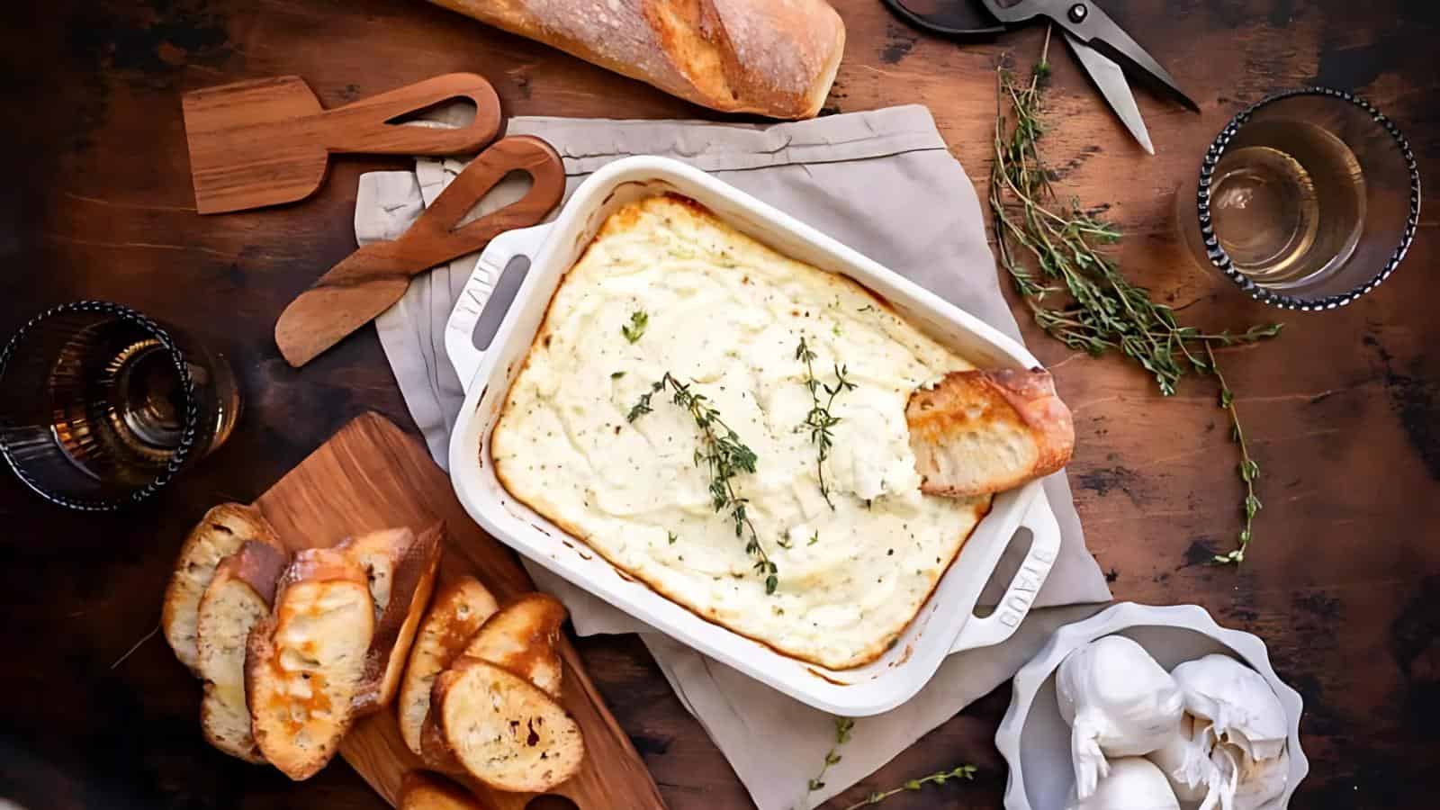 A dish of baked mashed potatoes topped with herbs, surrounded by slices of toasted bread, a loaf of bread, garlic bulbs, and a glass of water on a wooden table.
