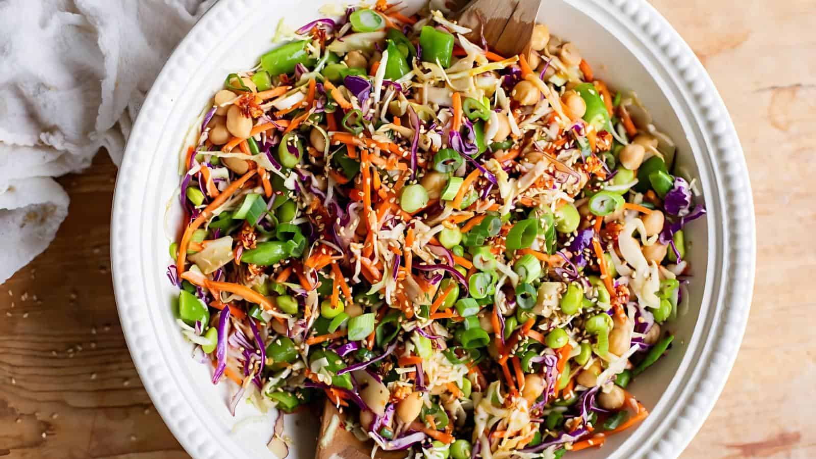 A large white bowl filled with a colorful salad containing shredded cabbage, carrots, snap peas, chickpeas, green onions, and sesame seeds, with wooden salad servers.