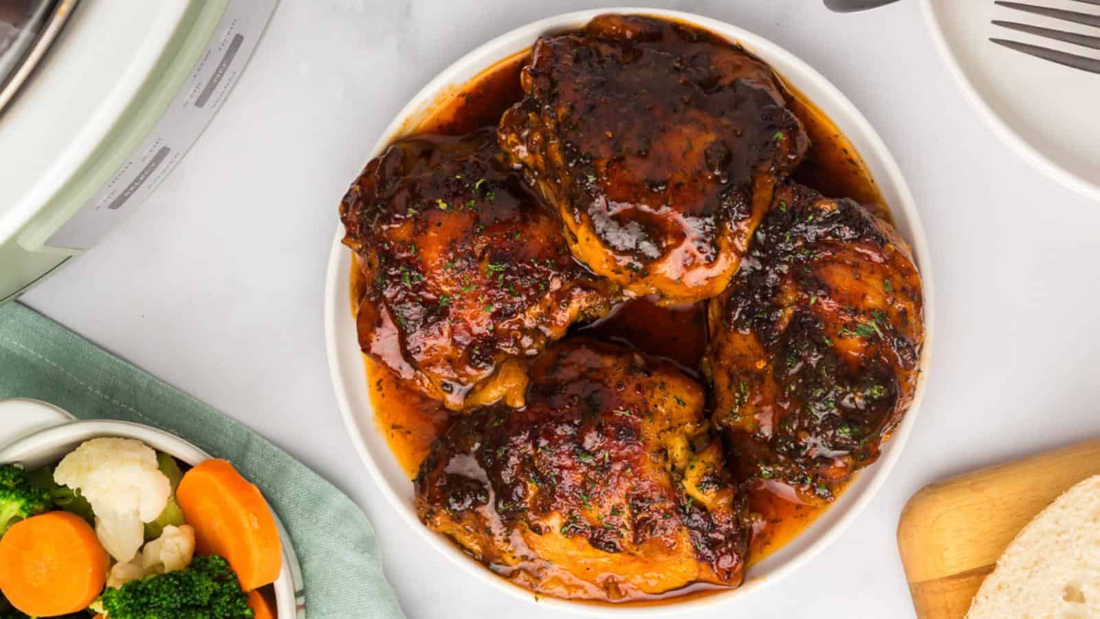 A white bowl filled with four glazed, cooked chicken thighs, surrounded by a bowl of mixed vegetables and a partial plate with bread.