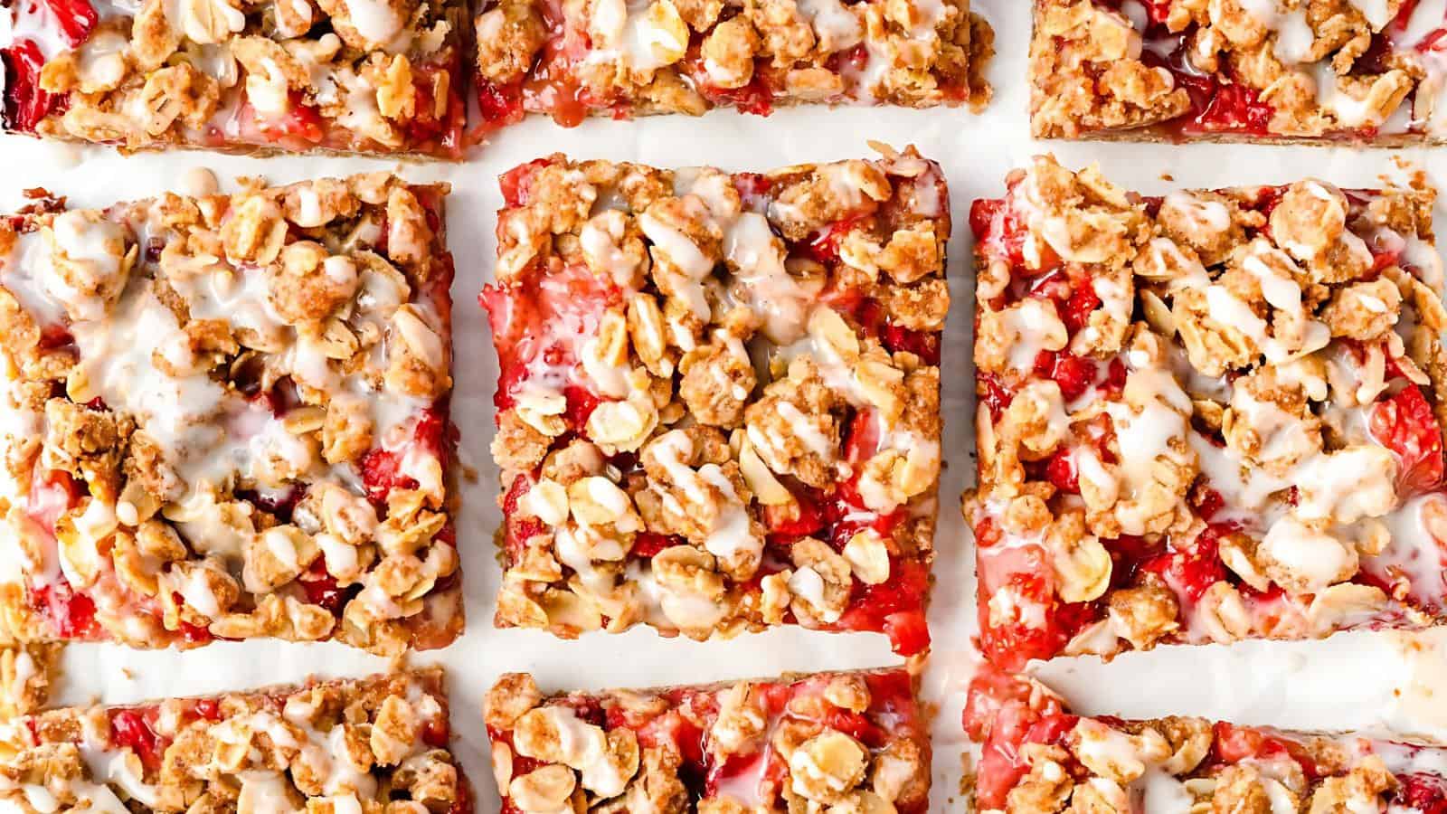 Nine strawberry oat crumble bars with white icing drizzle, arranged in a grid pattern on a white surface.