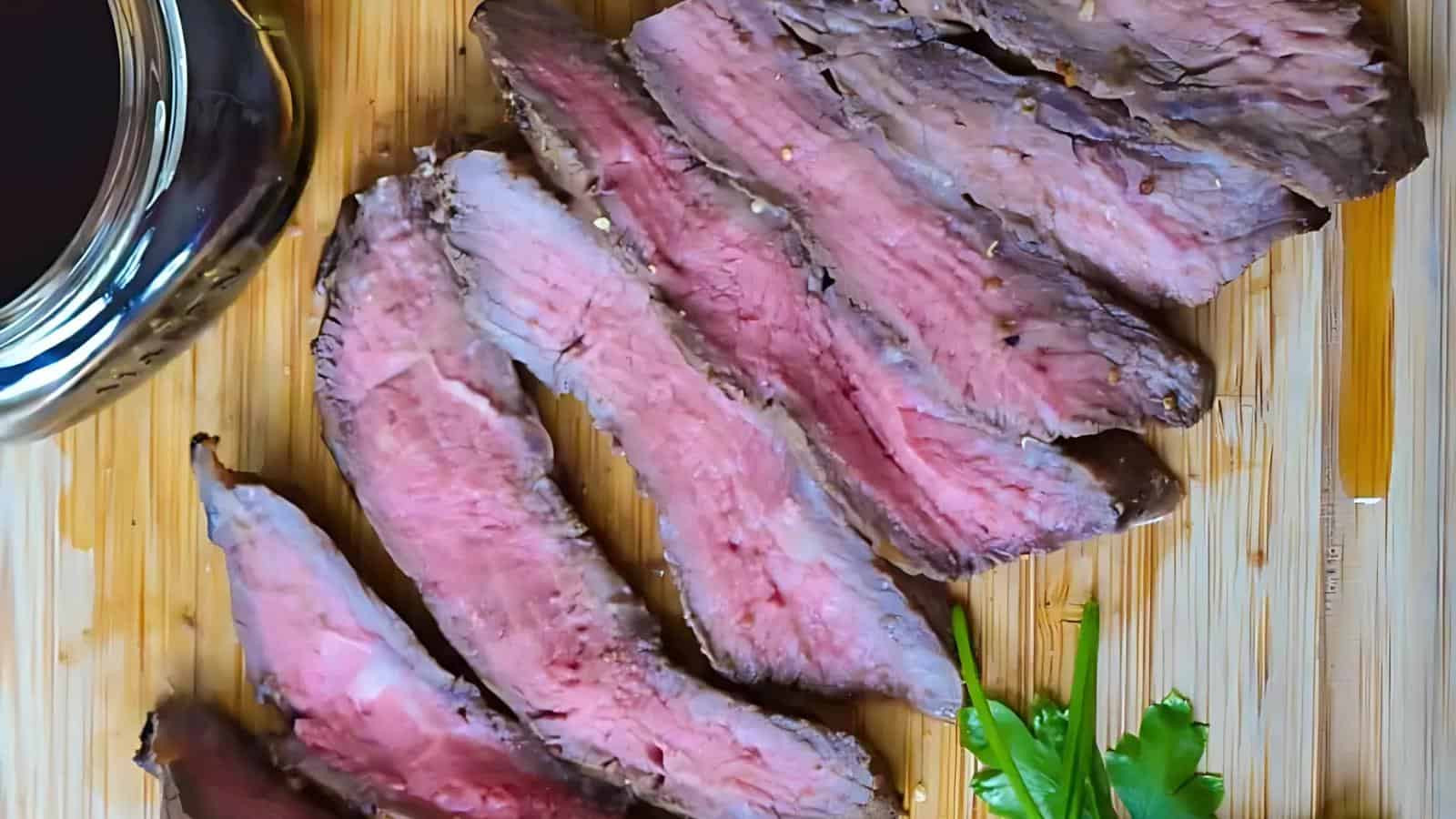 Sliced medium-rare steak arranged on a wooden cutting board with a glass of dark liquid and a sprig of parsley nearby.