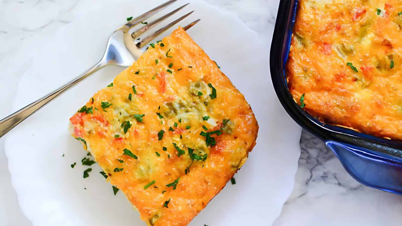 A slice of cheesy casserole with herbs, served on a white plate with a fork, next to a blue dish containing more casserole on a marble surface.