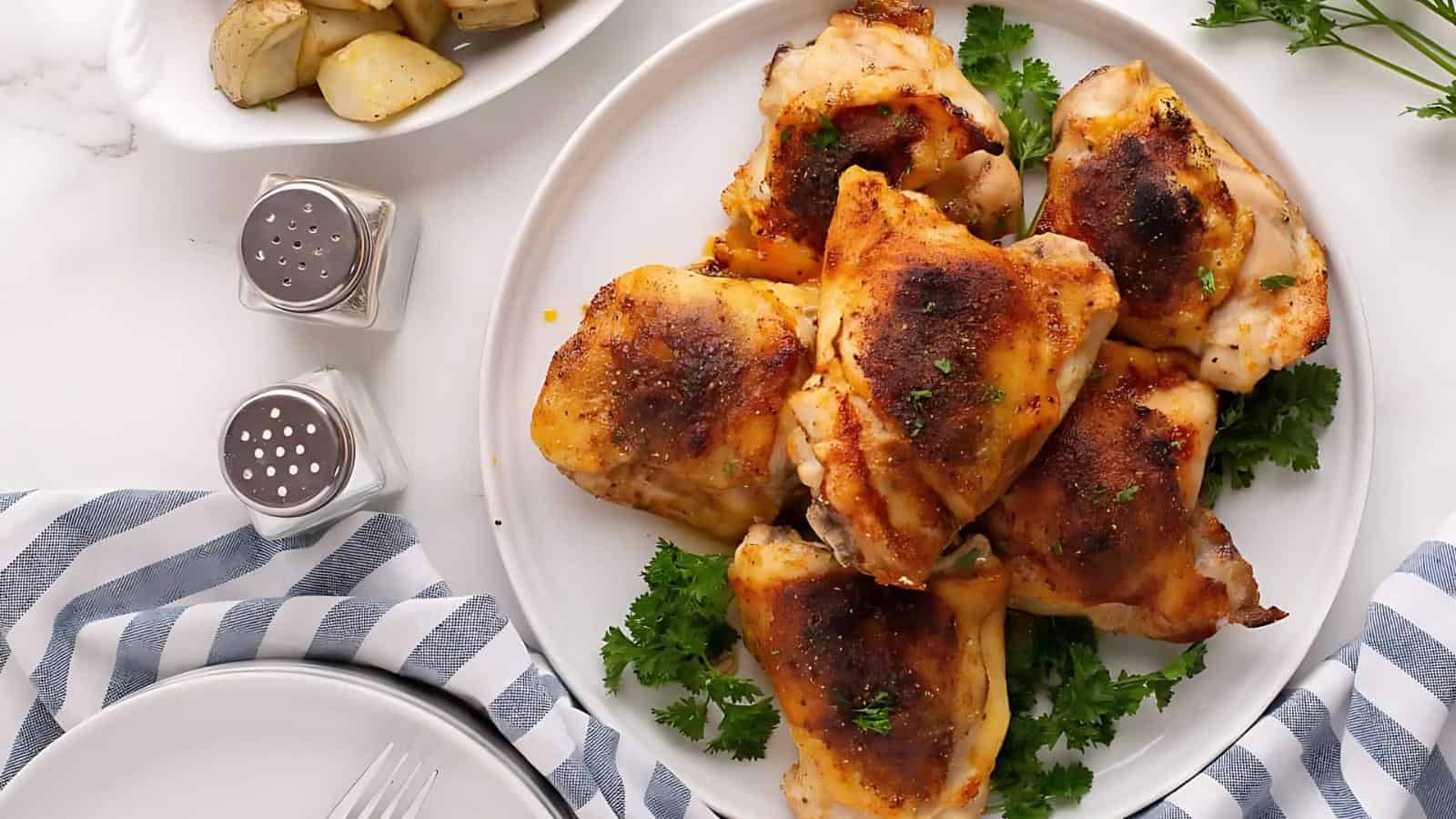 A plate of seasoned roasted chicken thighs on a bed of parsley, with salt and pepper shakers and a side dish of roasted potatoes nearby.