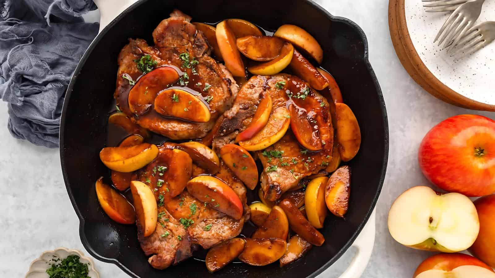 Pork chops in a cast iron skillet topped with sliced apples and sauce, garnished with herbs. A bowl of apples and a plate with forks are nearby.