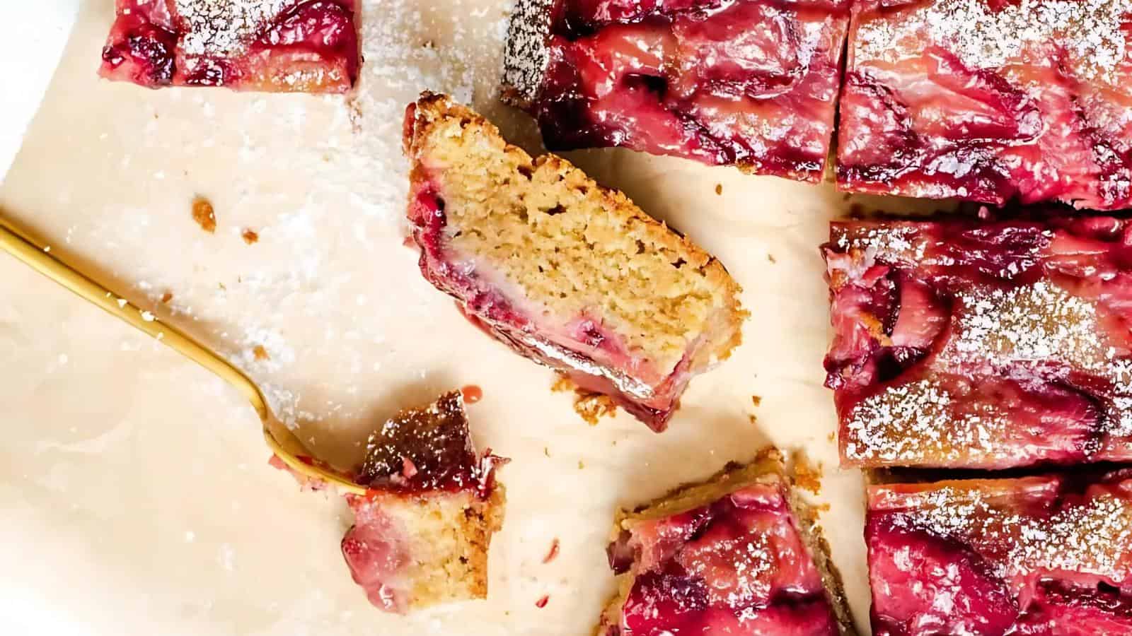 Sliced pieces of fruit-topped cake on parchment paper, with a fork holding one piece; cake appears moist with a dusting of powdered sugar.