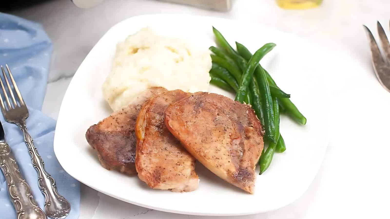 A plate with two cooked pork chops, mashed potatoes, and steamed green beans, set on a white surface with silverware and a blue napkin nearby.