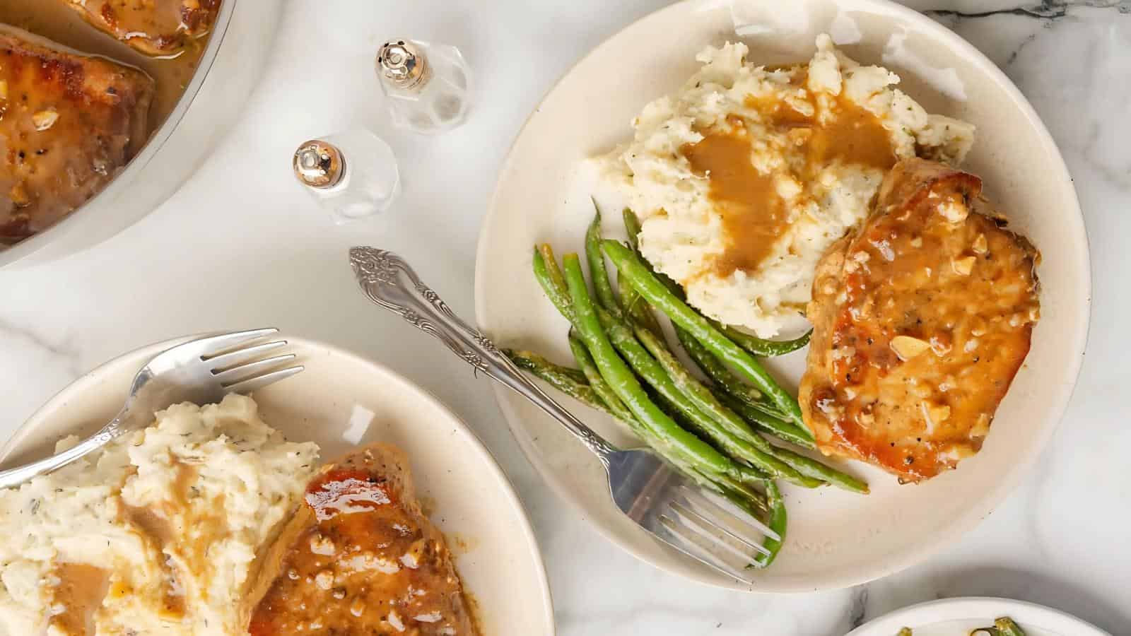 Two plates with mashed potatoes topped with gravy, green beans, and a grilled pork chop on a marble surface; forks and salt and pepper shakers are also visible.