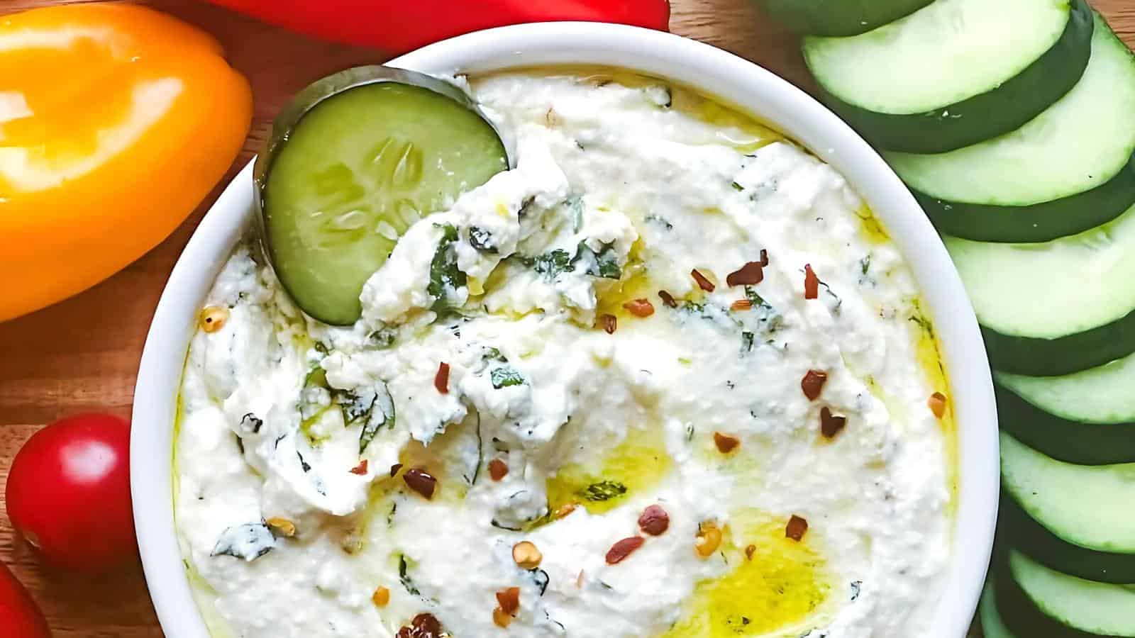 A bowl of herbed ricotta dip topped with olive oil and red pepper flakes, surrounded by slices of cucumber, yellow bell pepper, a cherry tomato, and a red bell pepper.