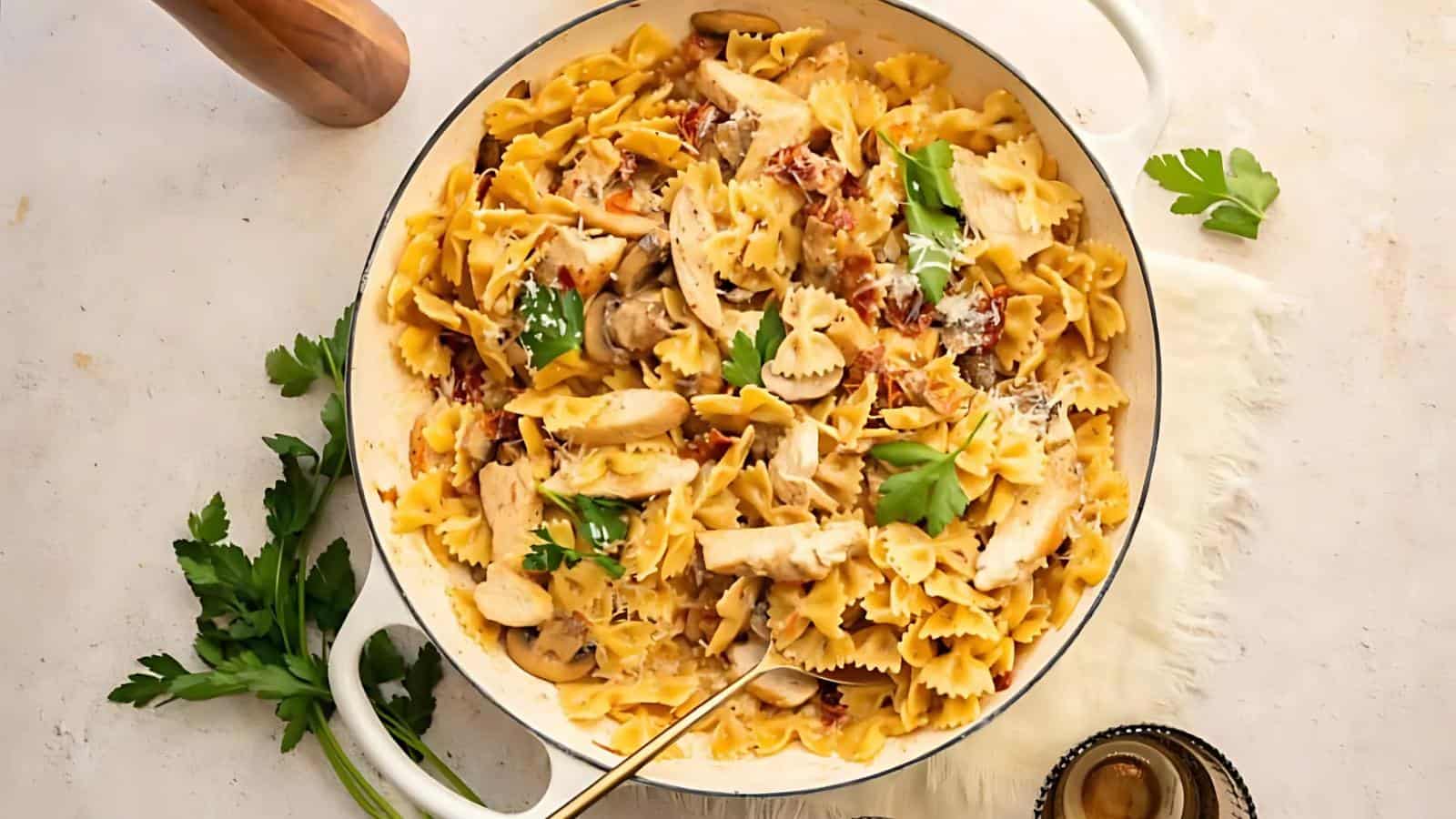 A pan of bowtie pasta with sliced chicken, mushrooms, sun-dried tomatoes, and fresh parsley, garnished with grated cheese.