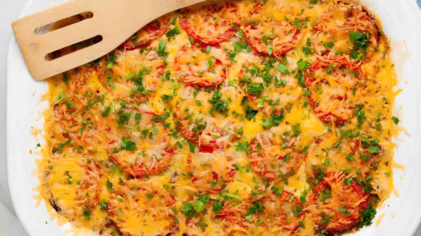 A baked casserole topped with melted cheese, sliced tomatoes, and chopped herbs, with a wooden spatula resting on the side.
