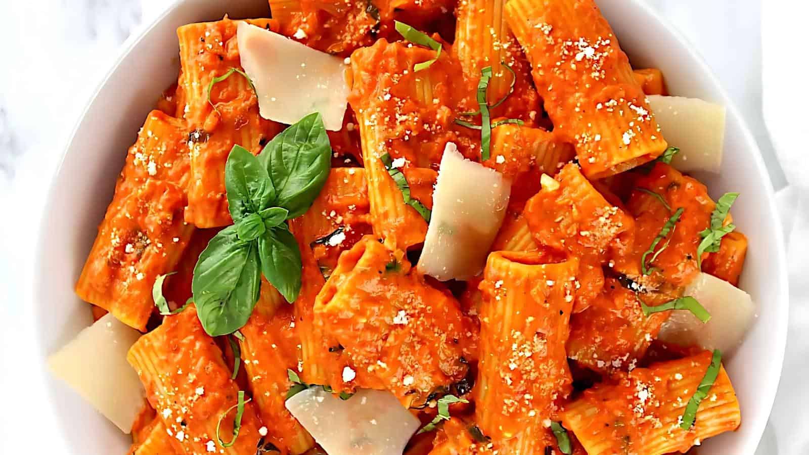 Bowl of rigatoni pasta in red sauce, topped with grated and shaved cheese, garnished with fresh basil leaves.