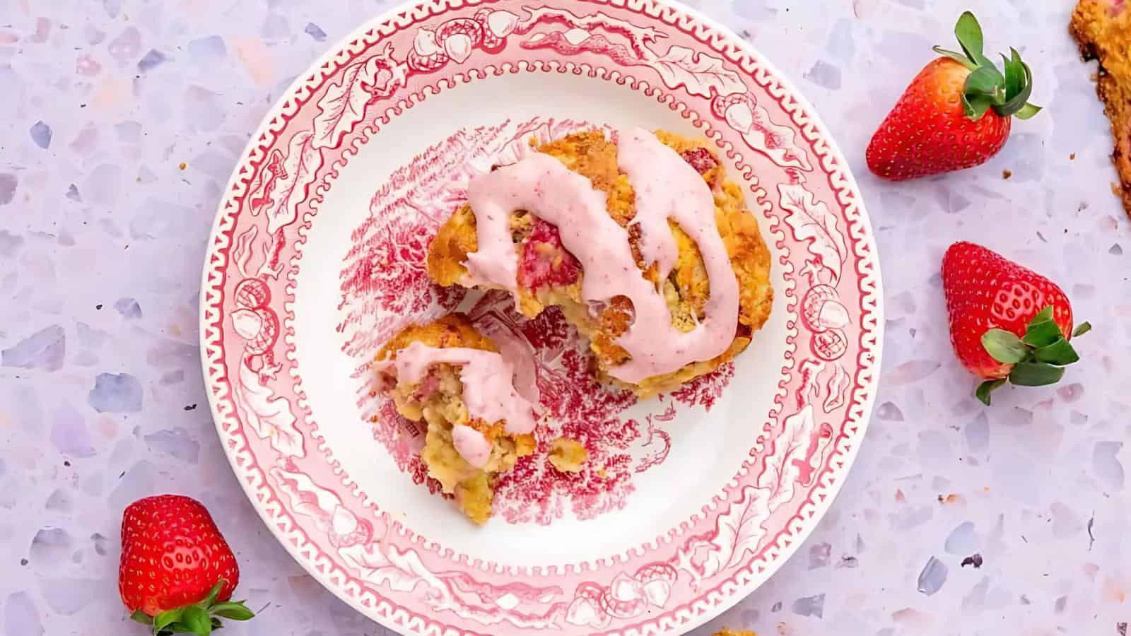 A partially eaten sweet roll with pink icing sits on a decorative plate, with fresh strawberries nearby on a speckled surface.
