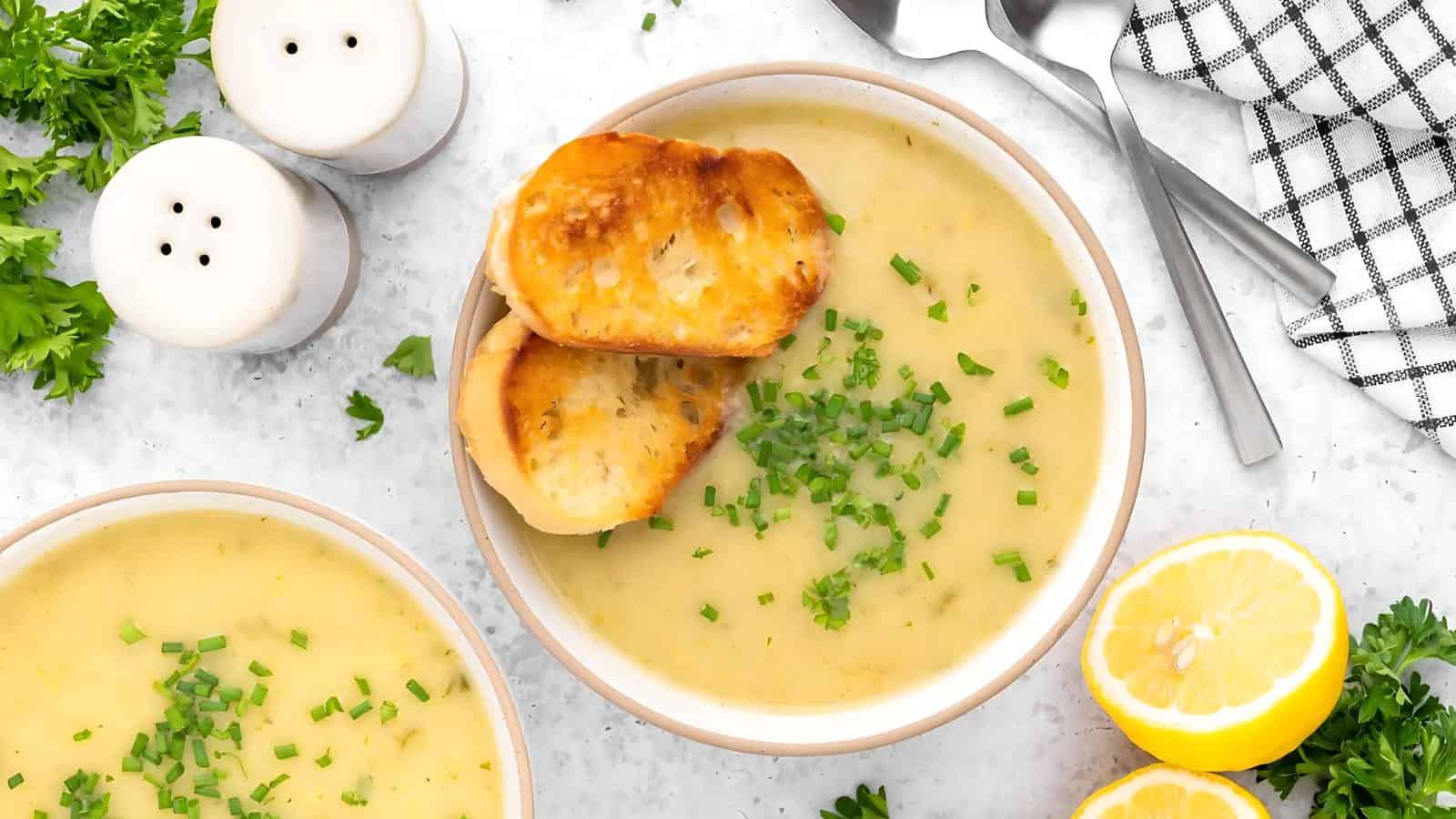 Bowl of creamy soup topped with chopped chives and two slices of toasted bread, surrounded by parsley, salt and pepper shakers, a lemon half, and utensils on a light surface.
