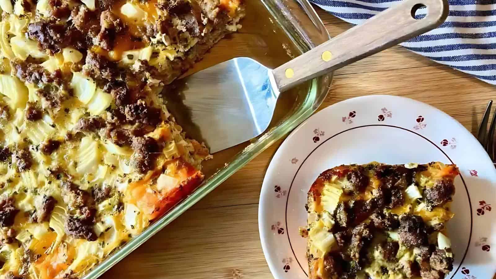 A baked casserole with ground meat and chopped vegetables in a glass dish. A square piece is served on a decorative plate beside the dish, with a spatula resting in the dish.