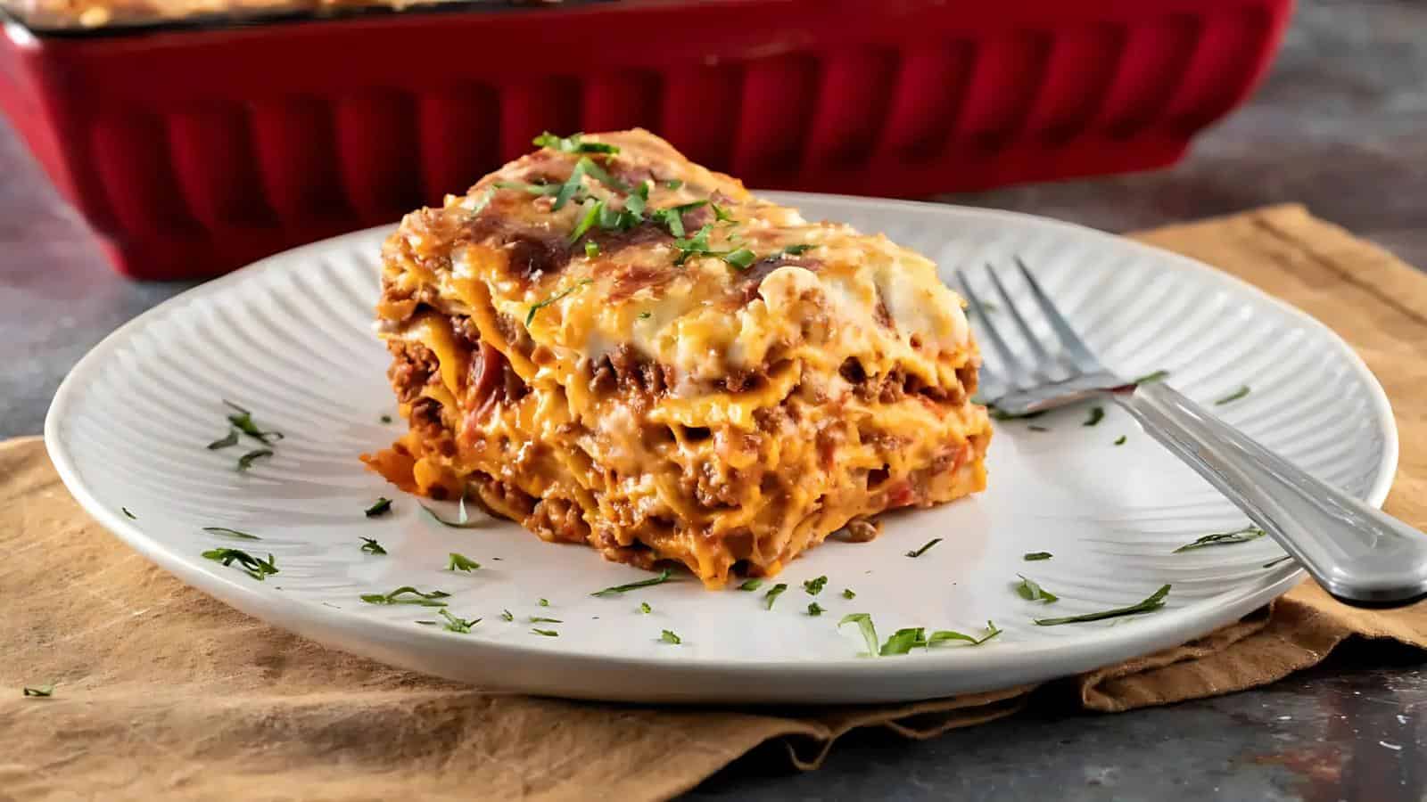 A slice of lasagna with meat and cheese sits on a white plate with a fork, garnished with chopped herbs. A red baking dish is in the background.